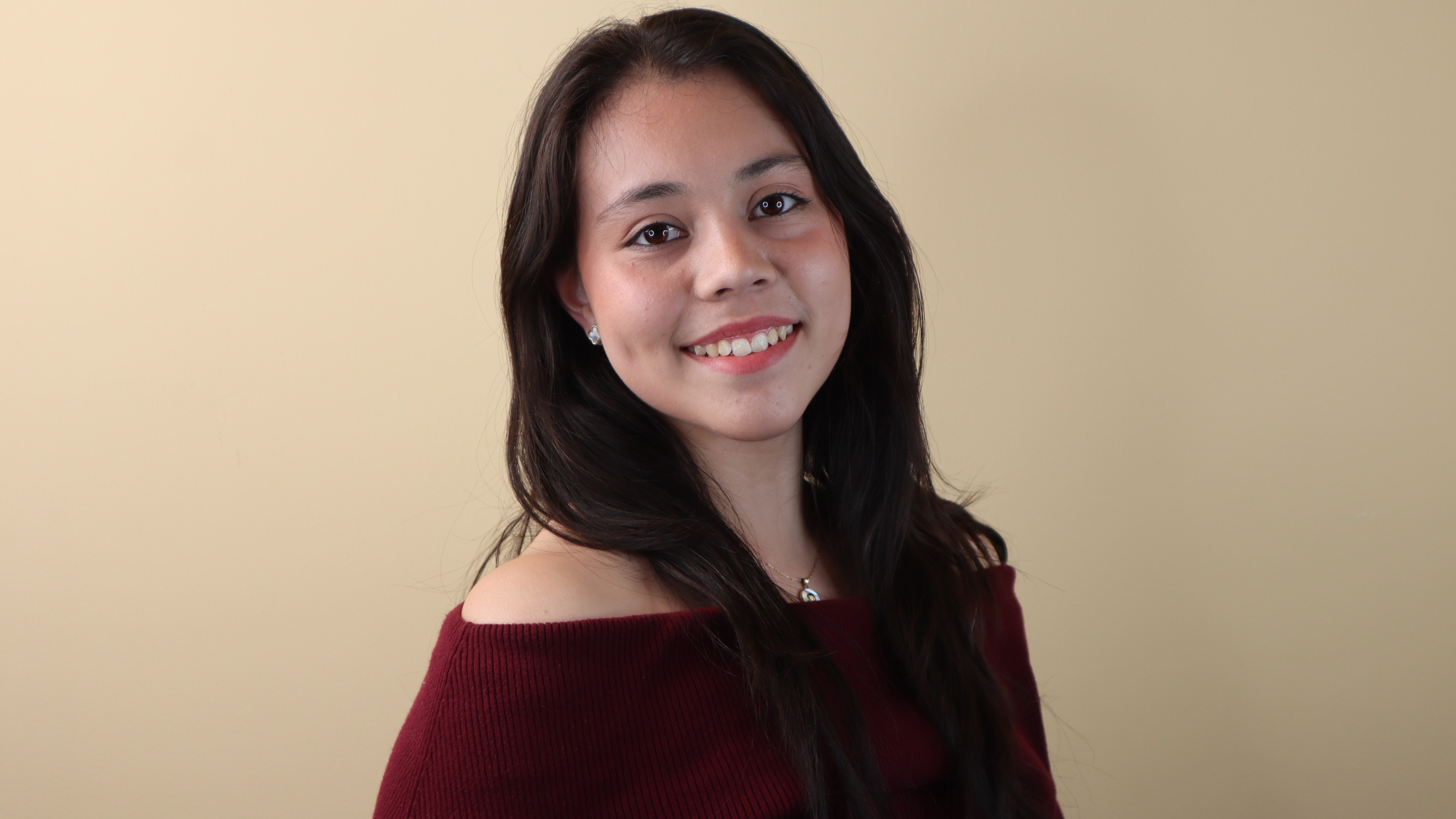 Maria Cortez Cruz smiles in front of a beige background while wearing an off-the-shoulder maroon sweater. Maria Cortez Cruz smiles in front of a beige background while wearing an off-the-shoulder maroon sweater.